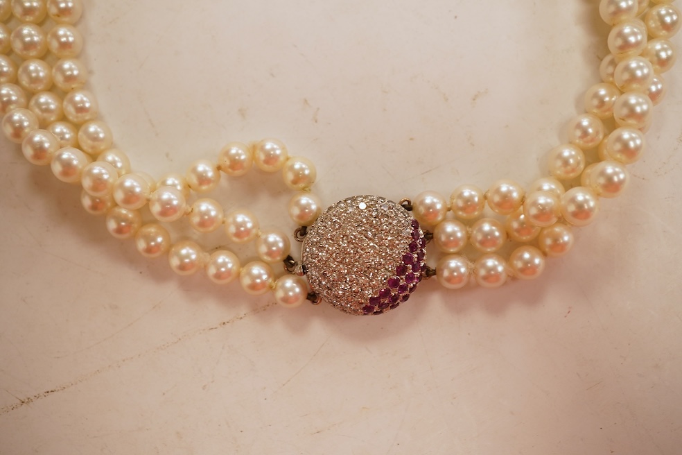 A cultured pearl, ruby and diamond clasp, composed of three rows of cultured pearls measuring approximately 6.9mm diameter, to a white precious metal (tests as 14k) disc-shaped clasp pavé-set with brilliant-cut diamonds